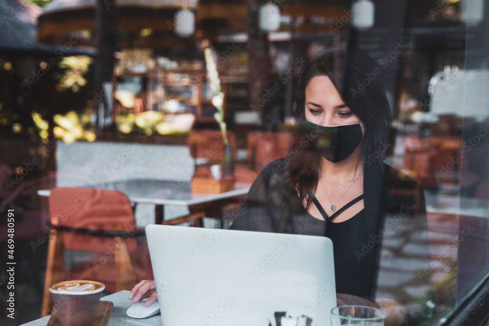 Young woman freelancer with protective medical black mask on face working with laptop in cafe