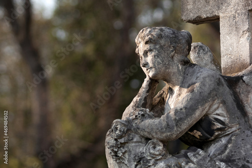 A sitting, pensive Angel. An angel with his head on his hand, looking at the sky. Blurred trees in the background. Stone, monumental sculpture in the Catholic cemetery.