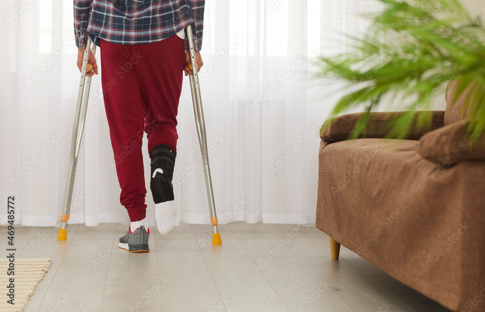 Unrecognizable man with broken foot or ankle walking on crutches. Patient wearing leg brace support fracture fixator standing by window in his apartment or rehabilitation center. Cropped low section Unrecognizable man with broken foot or ankle walking on crutches. Patient wearing leg brace support fracture fixator standing by window in his apartment or rehabilitation center. Cropped low section