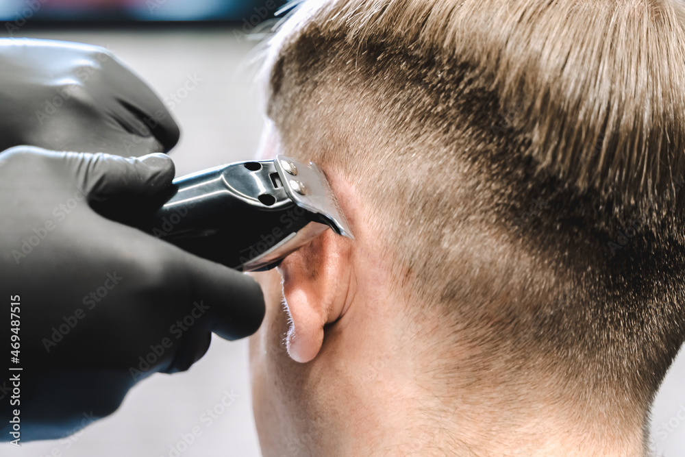 Visit to the barbershop. Hairdresser, barber shaves client's temples,sides with hair clipper. Young stylish man makes fashionable haircut.Customer sitting in dark men's beauty salon covered with cape Visit to the barbershop. Hairdresser, barber shaves client's temples,sides with hair clipper. Young stylish man makes fashionable haircut.Customer sitting in dark men's beauty salon covered with cape
