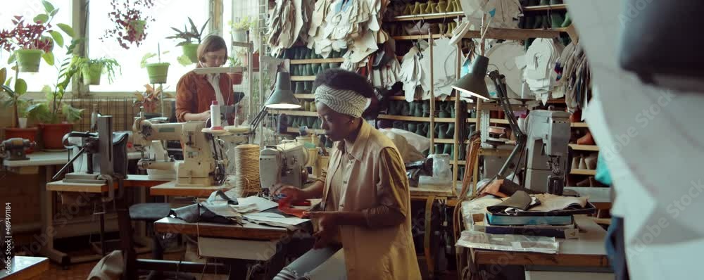Young African American female shoemaker working with leather at desk while her Caucasian colleague packing orders behind in workshop Young African American female shoemaker working with leather at desk while her Caucasian colleague packing orders behind in workshop