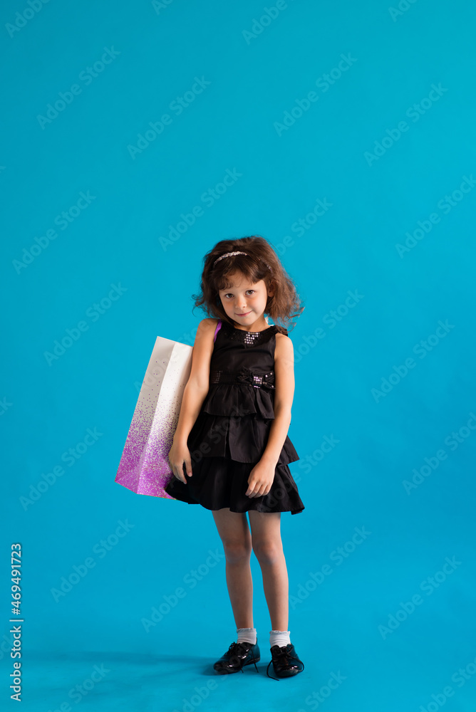 Cute cheerful girl in black dress with paper bags for shopping on blue background Cute cheerful girl in black dress with paper bags for shopping on blue background