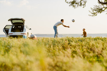 Young father and his son playing with ball by car at summer field