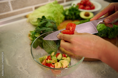 High angle view of female hands cutting vegetables with kitchen knife and putting them on glass transparent bowl. Vegan and healthy lifestyle concepts. Preparing raw vegan salad