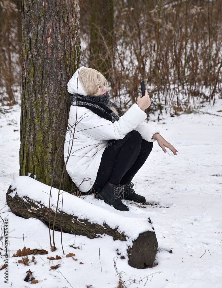 a girl wrapped in a scarf sits near a tree in winter and takes a selfie on her phone
