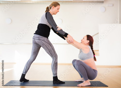 A pregnant woman is engaged in yoga. Exercise sitting on a roller  legs crossed and arms raised above them. A squatting pose  or a garland pose  or a malasana  holding the instructor s hands.