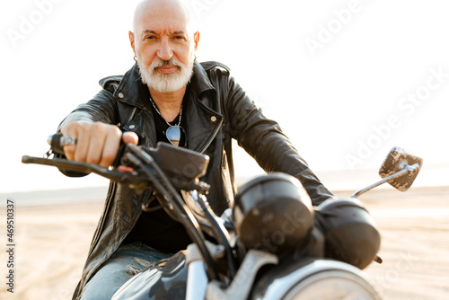 Mature bold man in leather jacket posing on motorbike