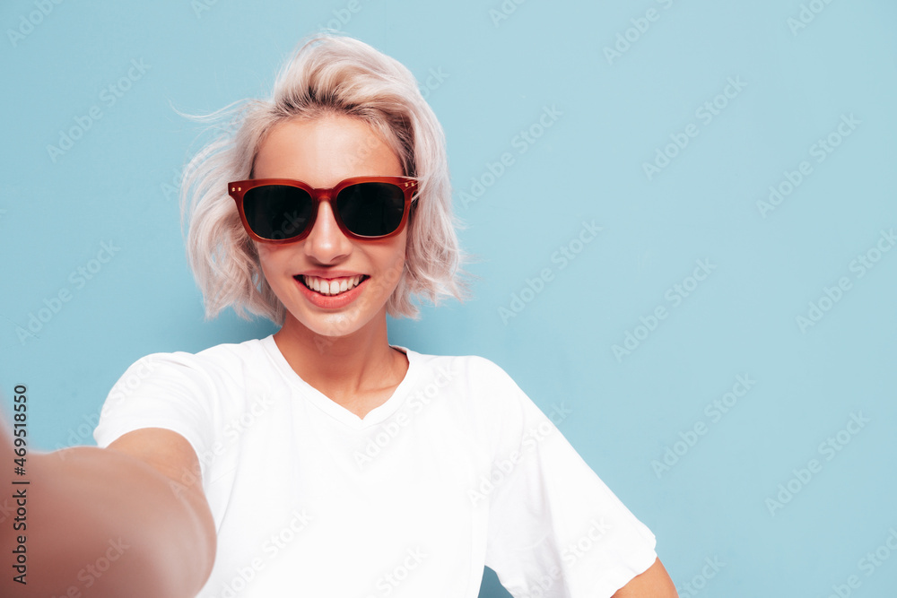 Young beautiful smiling female in trendy summer clothes. Sexy carefree woman posing near blue wall in studio. Positive blond model having fun in sunglasses. Cheerful and happy. Taking Pov selfie