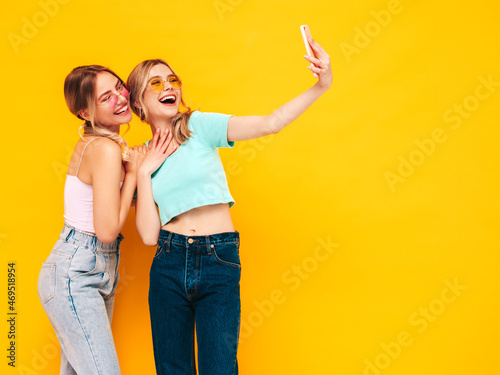 Two young beautiful smiling blond hipster female in trendy summer clothes. Sexy carefree women posing near yellow wall in studio. Positive models taking selfie. Cheerful and happy. In sunglasses