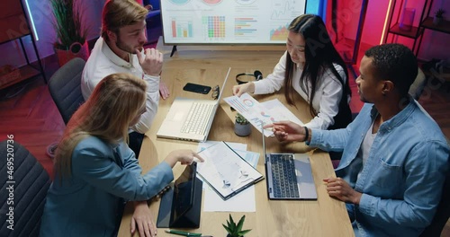 Top view of attractive confident experienced purposeful diverse team of men and women which sitting behind workplace and browsing paper reports and typing on computer in evening office