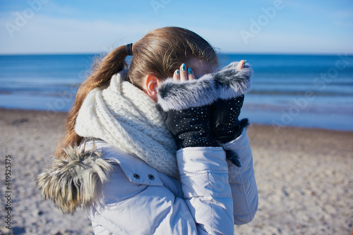 Dziewczyna spaceruje zimą nad Morzem Bałtyckim. Dłońmi w futrzanych rękawiczkach zasłania twarz przed fotografem.