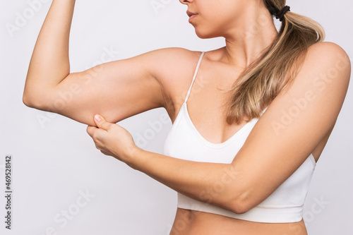 Cropped shot of a young caucasian blonde woman grabbing skin on her upper arm with excess fat isolated on a gray background. Pinching the loose and saggy muscles. Overweight concept photo