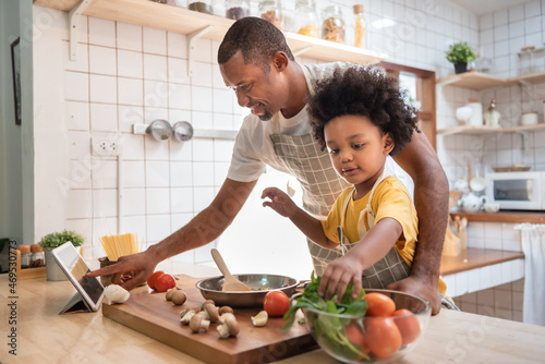 African American Little boy preparing food while his father looking on the digital recipe and using touch screen tablet in the kitchen