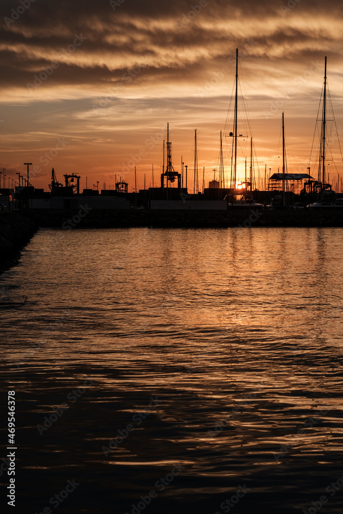 Sunset in the harbour with ships, masts and a lot of black sea with orange reflections.
