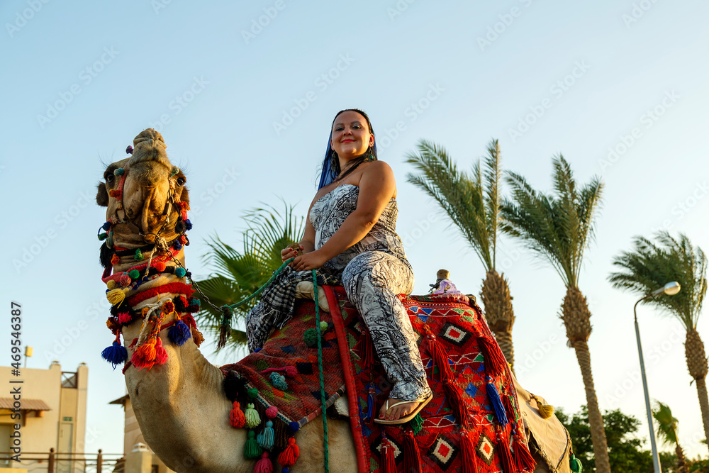 Woman tourist in a cape on her head rides a camel on a city street against the backdrop of palm trees.