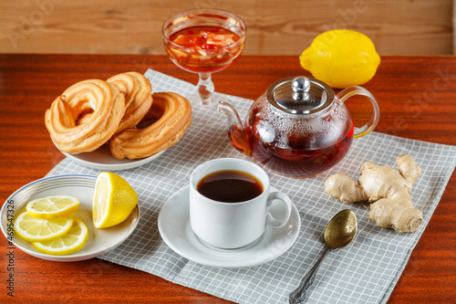 A teapot and a cup and saucer of strong aromatic tea on the table on a napkin next to ginger and lemon jam and donuts.