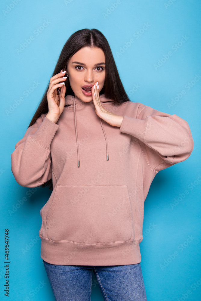 Brunette girl talking in surprise on the phone in a beige hoodie and jeans from behind blue background Brunette girl talking in surprise on the phone in a beige hoodie and jeans from behind blue background