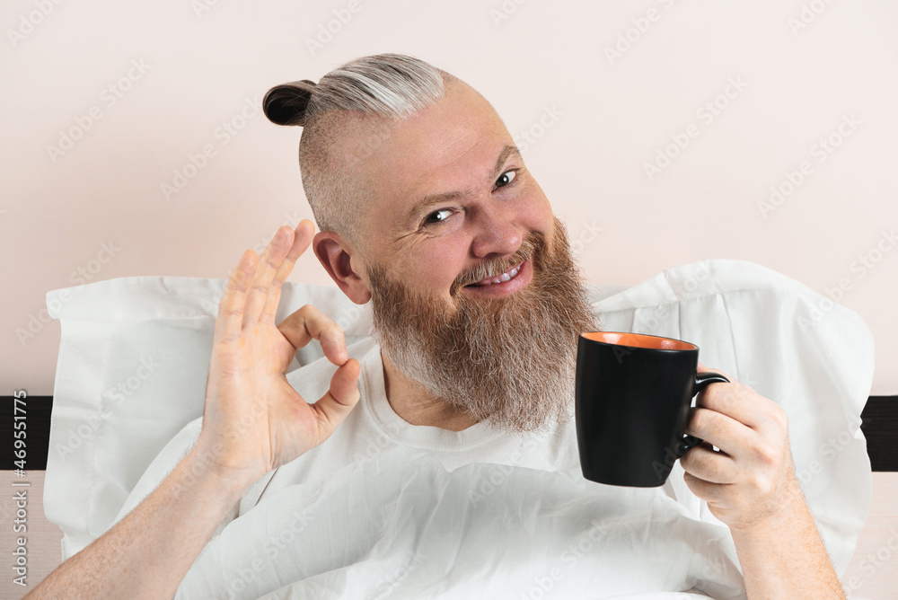 Happy bearded man enjoying morning coffee in bed showing OK gesture. Smiling hipster guy starting the day. Closeup