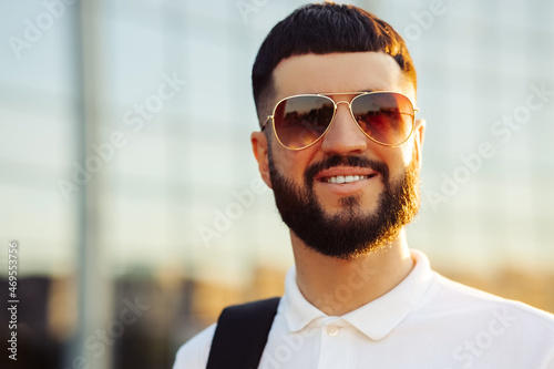 man with a beard in sunglasses, in a casual white shirt on a sunny day, smiles. Man outdoors near a glass building