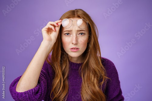 Waist-up shot of intense confused and unsure cute redhead female cannot look without glasses having bad sight taking off eyewear and squinting at camera uncertain, unable to read over purple wall photo