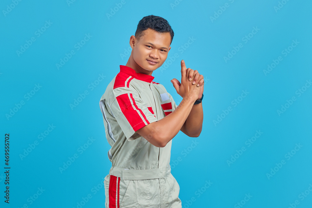 Portrait of young Asian mechanic holding symbolic weapon with hand gesture over blue background