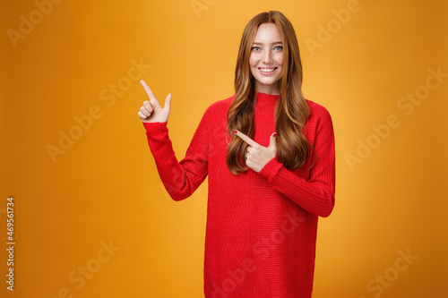 Assertive and helpful charming young female assitant in red knitted dress with ginger hair smiling friendly and pleasant as pointing at upper left corner, showing interesting thing over orange wall photo