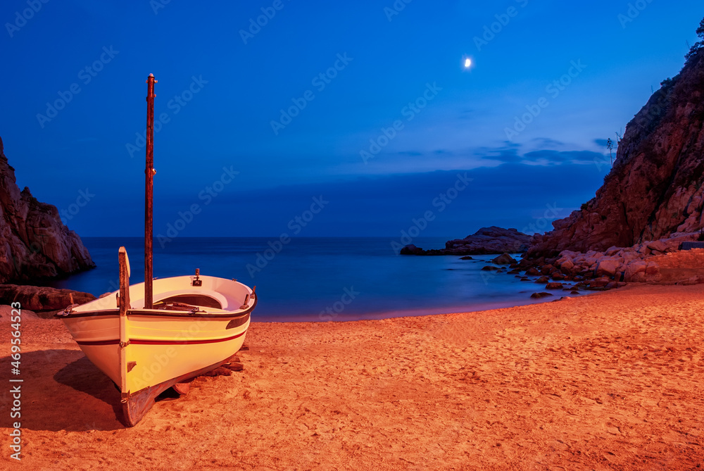 Naklejka premium A boat on a sandy beach in Tossa De Mar Bay lunar night against the backdrop of mountains and blue skies.
