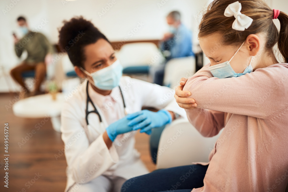 Small girl with face mask sneezes into elbow in waiting room at doctor's office. Small girl with face mask sneezes into elbow in waiting room at doctor's office.