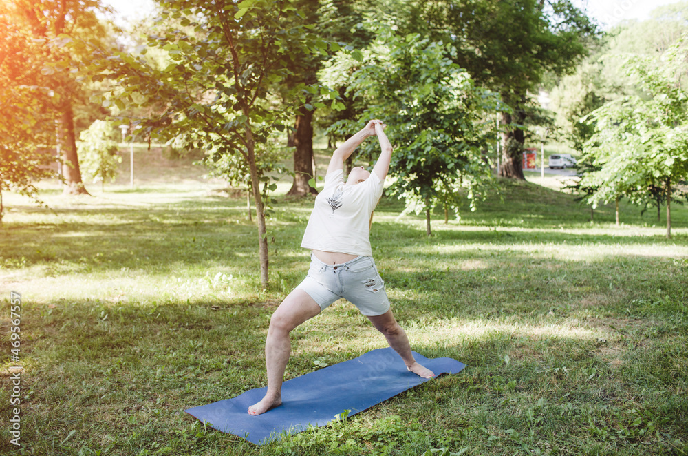 A mature woman in a T-shirt and shorts does fitness exercises and does yoga outdoors.