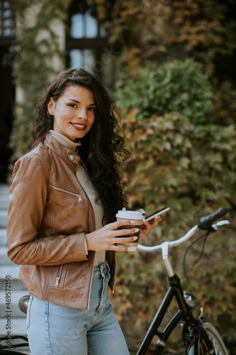 Young woman with mobile phone drink coffee to go by the bicycle on autumn day