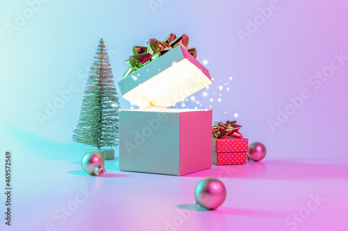 Neon Christmas winter background. Open xmas gift box with shine lights  minimal tree. Holiday decoration bauble ball on neon abstract gradient backdrop. Happy new year copy space.