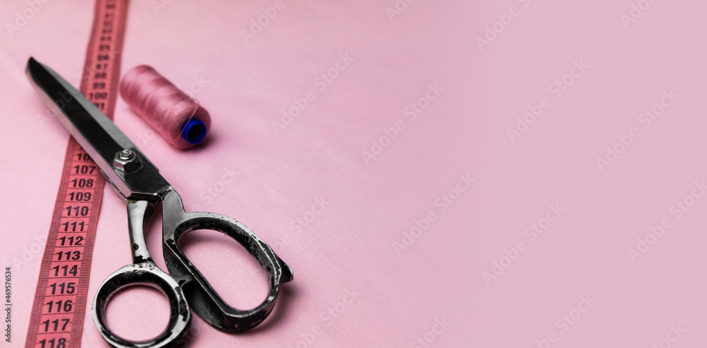 Fototapeta premium Spool of thread, centimeter and sewing scissors on a pastel pink background. Models for tailors or textile workshops. Empty space for text.