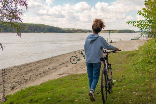 Young woman pushes a bicycle while walking along the river bank photo