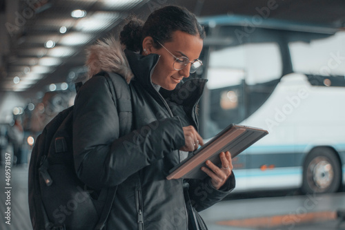 Turista empresaria con gafas formal con una sonrisa de pie revisando sus apuntes con contratos en la tableta digital en la estación de autobuses por el recinto para tomar apuntes del transporte