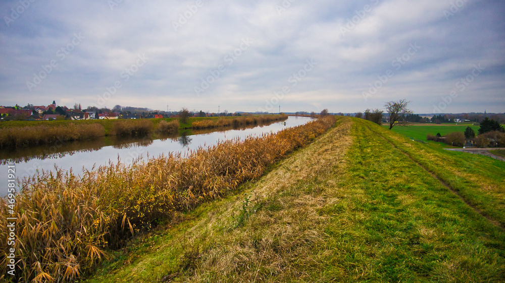 Obraz premium Blick entlang des Elster-Saale-Kanals mit herbstlichem Schilfufer und Deich unter bewölktem Himmel, Leipzig, Sachsen, Deutschland