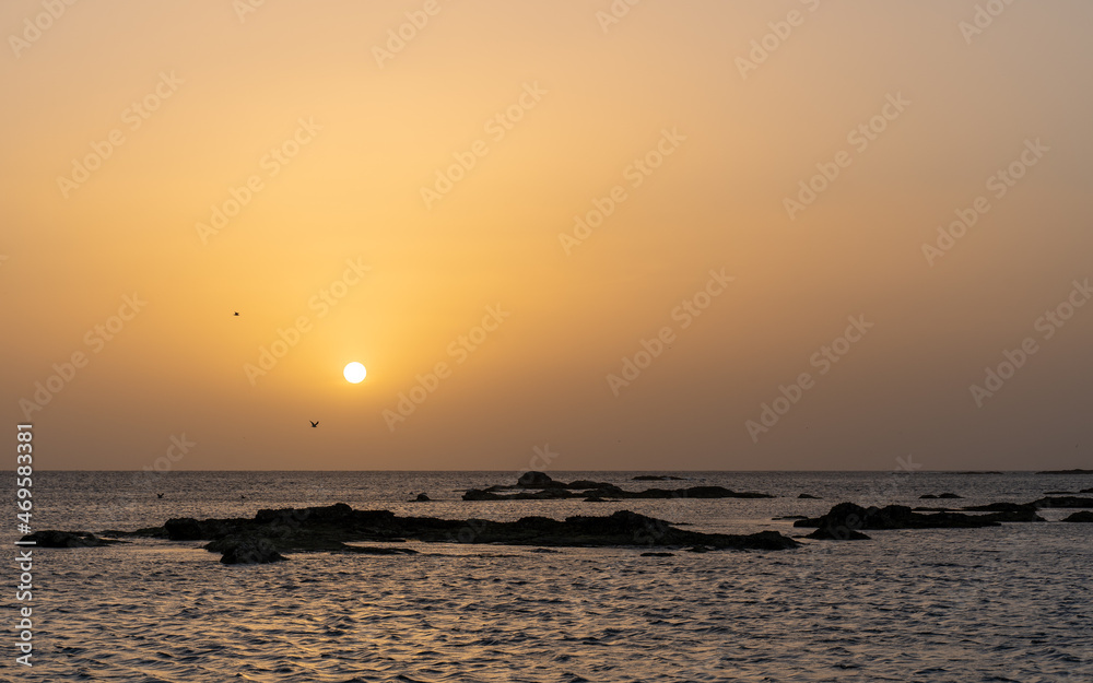 A tranquil Dhofar sunset, where the sun's descent casts a golden hue over the serene waters and rocky outcrops