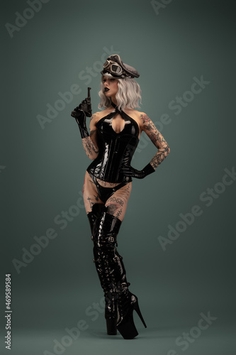 Pretty tattooed young woman wearing latex lingerie, cap and gun