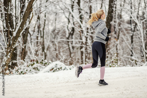 Rear view of fit slim sportswoman running in nature at snowy winter day. Winter fitness, cardio exercises, chilly weather