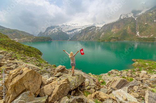 Tourist with swiss flag trekking by lakefront of Lake Robiei with dam. Swiss reservoir in Maggia Valley of Ticino canton. Top of aerial tramway station from San Carlo town of Switzerland.