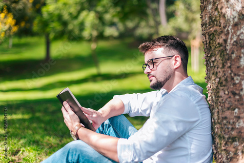 Young intelligent man wearing eyeglasses is reading book leaning on a tree in the city park