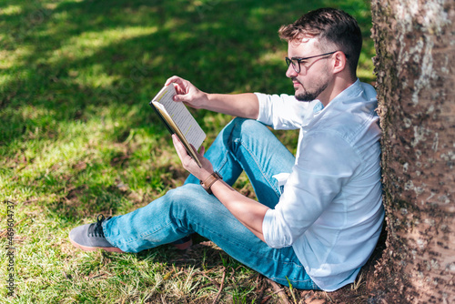 Young intelligent man wearing eyeglasses is reading book leaning on a tree in the city park
