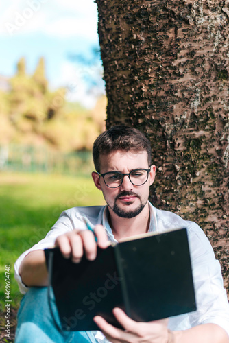 Young intelligent man wearing eyeglasses is reading book leaning on a tree in the city park