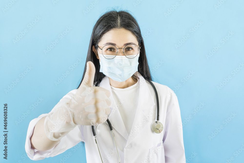 Young asian doctor woman wearing medical face mask,gloves , stethoscope over isolated blue background,thumbs up approving expression looking at the camera with showing success.