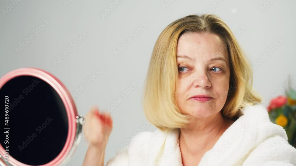 Mature woman combing blonde hair close-up, looking in mirror, using red comb. Beauty procedure, morning care routine in bathroom. Cosmetology concept, cosmetic products. Mature woman combing blonde hair close-up, looking in mirror, using red comb. Beauty procedure, morning care routine in bathroom. Cosmetology concept, cosmetic products.