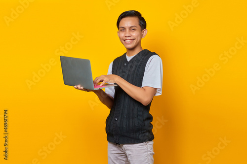 Smiling young handsome businessman holding a laptop and looking at camera on yellow background