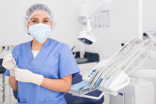 Positive female dentist in face mask standing near medical chair and preparing for treatment