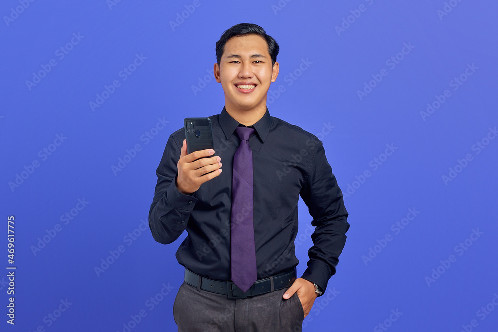 Cheerful young handsome businessman holding smartphone and looking at camera on purple background Cheerful young handsome businessman holding smartphone and looking at camera on purple background