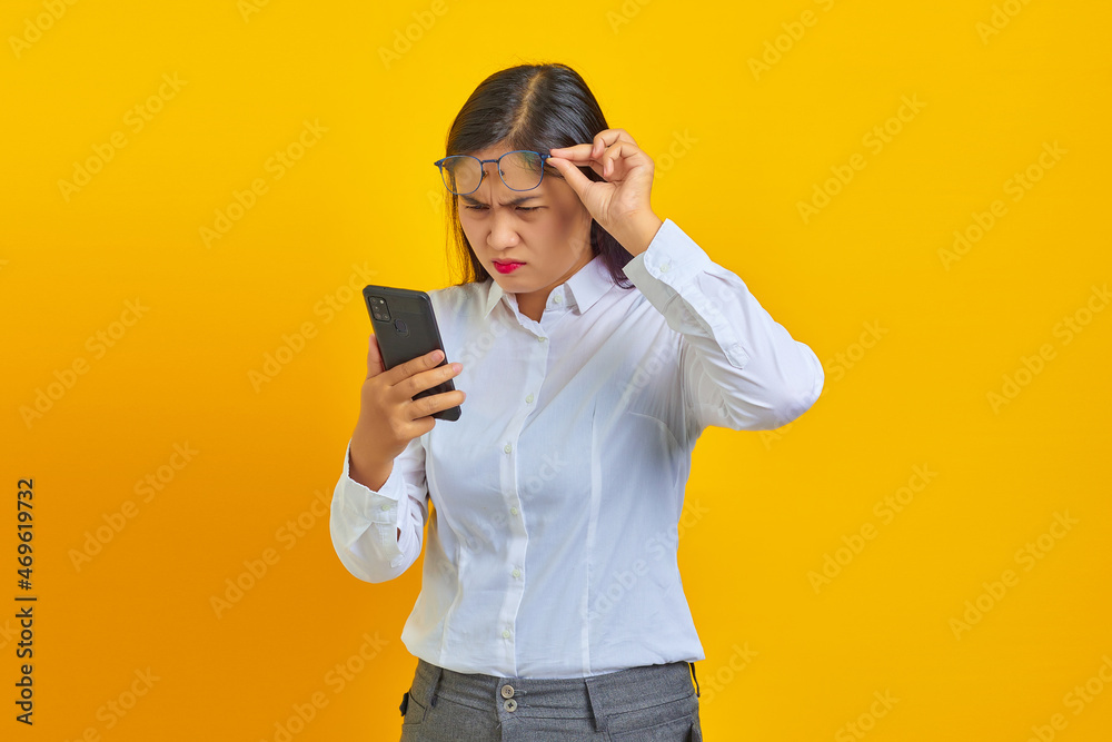 Angry young asian woman using mobile phone and taking glasses isolated over yellow background Angry young asian woman using mobile phone and taking glasses isolated over yellow background