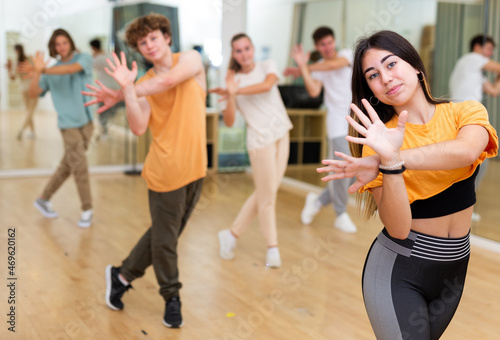 Happy brunette teenage girl dancer practicing active vigorous dance with group in modern studio.