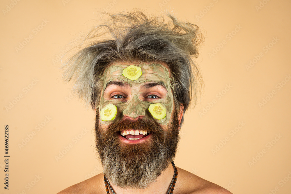 Funny man closeup with clay mask and cucumber slices on face. Spa, dermatology, wellness and facial treatment concept. Man having cosmetic moisturizing mask isolated studio background. Male face care.